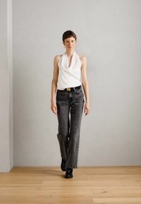Hvit halterneck-top med samling detaljer, kombinert med grå høytlivsjeans. Modellen har på seg svarte ankelboots og står på tregulv.