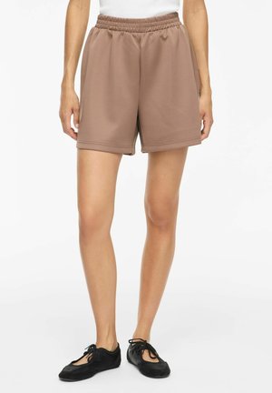 Personne portant un short marron clair à taille élastique et des chaussures plates noires à lacets, debout sur un fond blanc.
