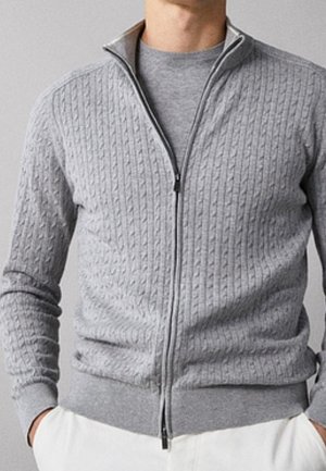 Mężczyzna ubrany w jasnoszary sweter zapinany na zamek z wzorem warkoczy, pod którym ma szarą koszulkę z okrągłym dekoltem, ręce w kieszeniach, noszący białe spodnie.