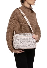 Borsa a tracolla beige con motivo e dettagli del logo, forma rettangolare, superficie testurizzata e tracolla regolabile, tenuta da una persona in un maglione di lana.
