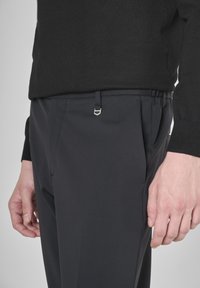 Persona con pantaloni neri e un maglione nero, che mostra la tasca laterale e un piccolo dettaglio metallico vicino alla vita.