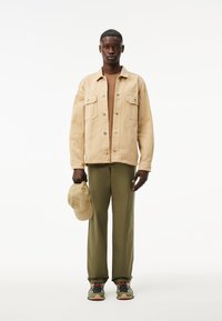 Giacca di denim beige con due tasche sul petto, pantaloni verde oliva, cappellino beige in mano e sneakers grigie con accentuazioni di colore. Sfondo semplice.