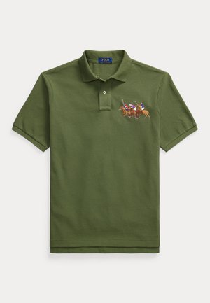 Homme portant un polo vert olive avec une broderie de cavaliers, un pantalon plissé blanc, des mocassins à pampilles marron et une montre-bracelet, debout devant un fond neutre.