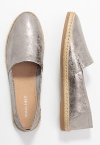 Par de espadrilles metálicas plateadas de tipo slip-on con adornos de yute trenzado y suelas de goma marrón claro, mostradas desde vistas superior y lateral.