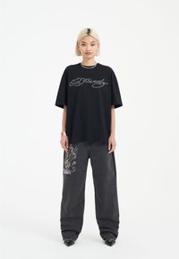 Camiseta negra de gran tamaño con el logo "Ed Hardy" en plata. Combinada con unos pantalones vaqueros de pierna ancha negros que presentan un diseño bordado colorido en la pierna.