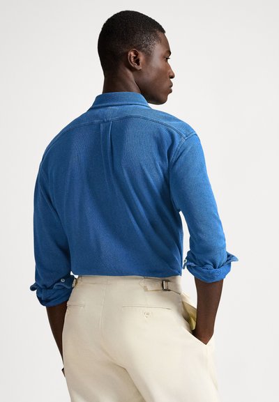 Mėlyna, ilgomis rankovėmis marškiniai su atsegama apykakle ir užlenktais rankogaliais, derinami su šviesiai smėlio spalvos kelnėmis, turinčiomis galines kišenes ir diržo detalę.