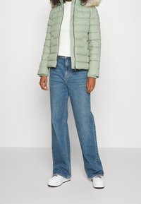 Miętowa pikowana kurtka z kapturem wyłożonym futrem, połączona z jasnoniebieskimi dżinsami o szerokich nogawkach i białymi trampkami. Prosty, funkcjonalny design.