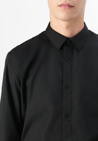 Homme portant une chemise noire à col boutonné, cadré du cou jusqu'en haut de la poitrine, sur un fond blanc uni.