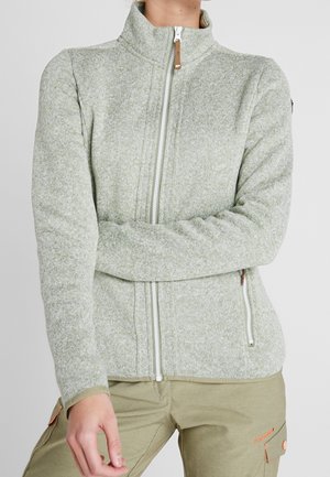 Osoba ubrana w jasnoszarą polarową kurtkę na zamek z kieszeniami typu kangur oraz beżowe spodnie z pomarańczowymi szwami.