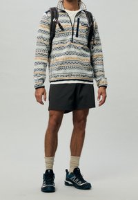 Polaire à enfiler bleu et crème avec des motifs en zigzag, poche poitrine zippée, associée à un short gris et des chaussures de randonnée bleu sarcelle.