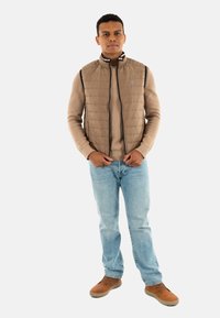 Gilet rembourré marron clair avec col montant sur un pull beige, associé à un jean bleu clair et des chaussures marron. Design simple, sans motifs visibles.