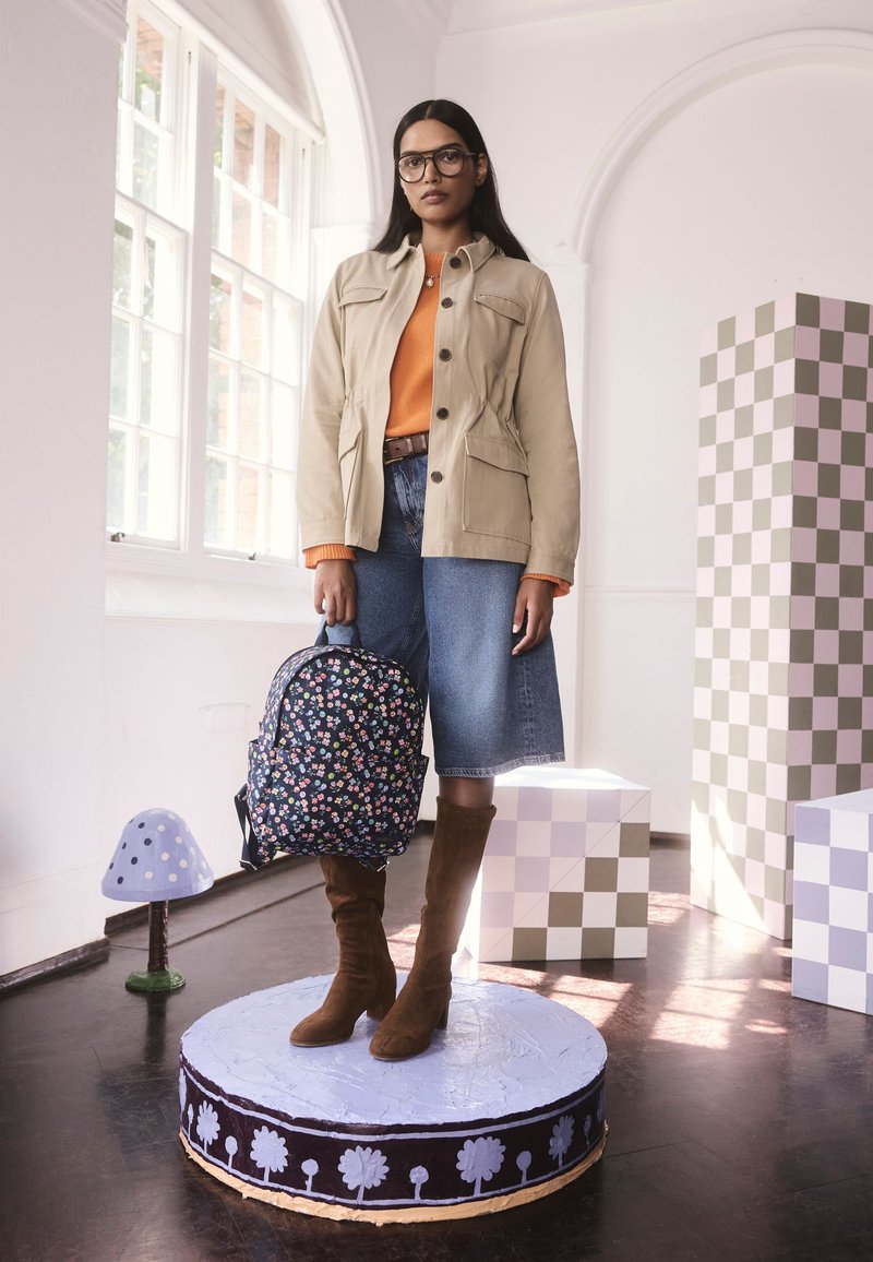 Bézs vászon dzseki, narancssárga pulóver, farmer szoknya, barna velúr térdig érő csizma, virágos mintás hátizsák, lila köralakú talapzaton állva.