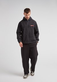 Giacca nera con zip e cappuccio, con un logo rosso sul petto, realizzata in tessuto liscio e dotata di tasche laterali, abbinata a pantaloni neri larghi.