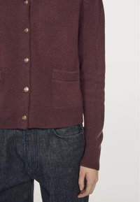 Burgundy knitted cardigan with long sleeves, front pockets, and gold buttons. Soft texture with ribbed cuffs and hem, paired with dark denim.
