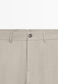 Pantalon beige clair avec fermeture à bouton, passants pour ceinture et braguette, présenté de la taille au milieu de la cuisse.