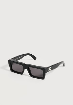 Lunettes de soleil rectangulaires noires avec des montures épaisses et des verres foncés posées sur une surface blanche, avec un petit logo blanc sur les branches.