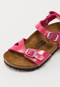Rosa lackläder sandal med justerbara silver spännen, korkfotbädd och svart gummisula. Har korsade remmar och en platt design.