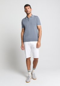 Grijze polo met korte mouwen en witte rand, gedragen met witte shorts en grijze sneakers. Gestreepte groen-witte sokken zichtbaar.