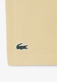 Tela beige con un pequeño logo de cocodrilo verde bordado cerca del borde inferior, mirando hacia la derecha con la boca abierta.