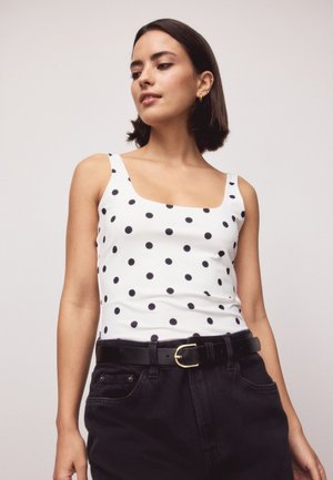 Jeune femme aux cheveux courts et foncés portant un haut blanc sans manches à pois noirs et un pantalon noir taille haute avec une ceinture.