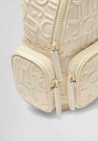 Zaino color crema con motivo del logo in rilievo, superficie testurizzata, doppie zip e tasca inferiore arrotondata per ulteriore spazio di stoccaggio.
