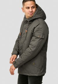 Olive green insulated jacket with a hood, drawstring, side pockets, and button accents. Features a zippered pocket on the sleeve.