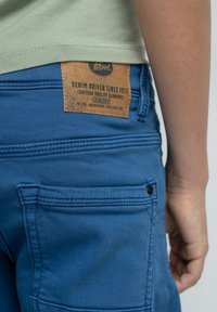 Los pantalones de mezclilla azul presentan un acabado texturizado con múltiples bolsillos. Una etiqueta marrón en la parte posterior muestra la marca y los detalles de cuidado.