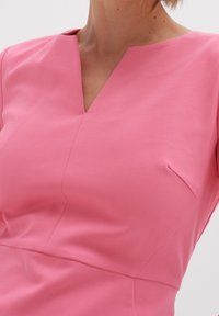 Rosa klänning med V-ringning, korta ärmar och figurnära livstycke. Har en diskret sidficka och mjukt, stretchigt tyg.