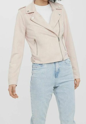 Woman wearing pale pink cropped biker jacket with silver zippers over white shirt and light blue high-waisted jeans on a plain background.