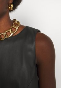 Top senza maniche in pelle nera con una texture liscia e un colletto rotondo, abbinato a una collana di catena dorata e orecchini a cerchio.