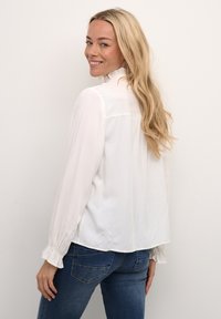 Weiße Bluse mit hohem Kragen und langen Ärmeln, ausgestattet mit rüschierten Manschetten und einer leicht lockeren Passform, kombiniert mit dunkelblauen Jeans.