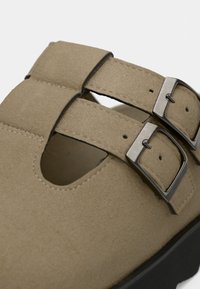 Μπεζ σουέντ παπούτσι με διπλούς ρυθμιζόμενους μεταλλικούς αγκράφες, σχεδιασμό με ανοιχτά κοψίματα, υφή στην επιφάνεια και μαύρη λαστιχένια σόλα.