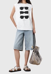 Camiseta sin mangas blanca con un gráfico de tres pares de gafas de sol negras, combinada con pantalones cortos de mezclilla azul claro y sandalias metálicas.