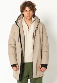 Hellbeige gepolsterte Jacke mit einer Kapuze, die schwarze Akzente, einen Reißverschluss und elastische Bündchen aufweist. Trägt man über einem cremefarbenen Pullover und grünen Hosen.