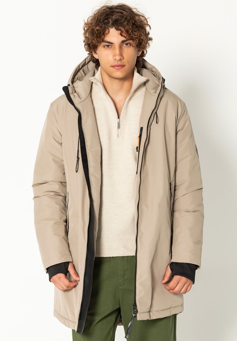 Hellbeige gepolsterte Jacke mit einer Kapuze, die schwarze Akzente, einen Reißverschluss und elastische Bündchen aufweist. Trägt man über einem cremefarbenen Pullover und grünen Hosen.