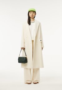 Lungo cappotto di cashmere crema, maglione bianco, pantaloni crema chiara, berretto verde e borsa a tracolla trapuntata verde scuro. Le scarpe casual completano l'outfit.