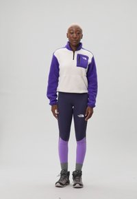 Jachetă din fleece în violet și alb, cu fermoar pe jumătate și buzunar. Asortată cu leggings negri și violet și șosete gri, completată cu bocanci de drumeție.