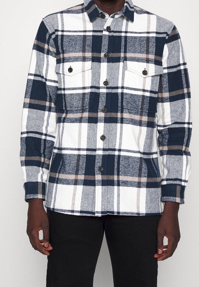 Homme portant une chemise à manches longues boutonnée avec un motif à carreaux bleu marine, blanc et beige et deux poches poitrine avant boutonnées.