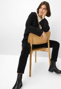 Frau in schwarzem Hoodie, schwarzen Hosen und schwarzen Stiefeln, die rückwärts auf einem Holzstuhl sitzt, ihren Kopf auf ihrer Hand stützt und zur Seite schaut.