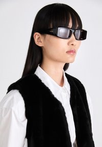 Schwarze rechteckige Sonnenbrille mit glatter Oberfläche, versehen mit einem dezenten Markensymbol, getragen von einem Model in einer schwarzen Kunstfellweste über einem weißen Hemd.