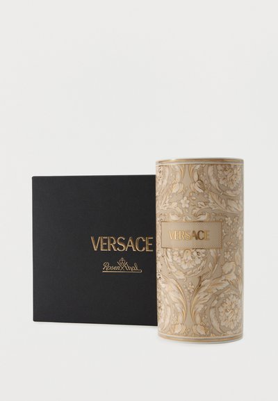 Boîte Versace carrée noire avec lettrage doré à côté d'un contenant cylindrique beige et doré orné de la marque Versace.