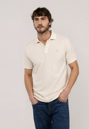 Hombre con cabello corto y rizado y barba, vestido con una camiseta tipo polo beige de manga corta y vaqueros azules, de pie con las manos en los bolsillos contra un fondo liso.