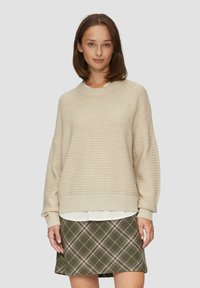 Beige gerippter Pullover mit rundem Halsausschnitt und überschnittenen Schultern, über einem weißen Hemd getragen, kombiniert mit einem grün karierten Rock.