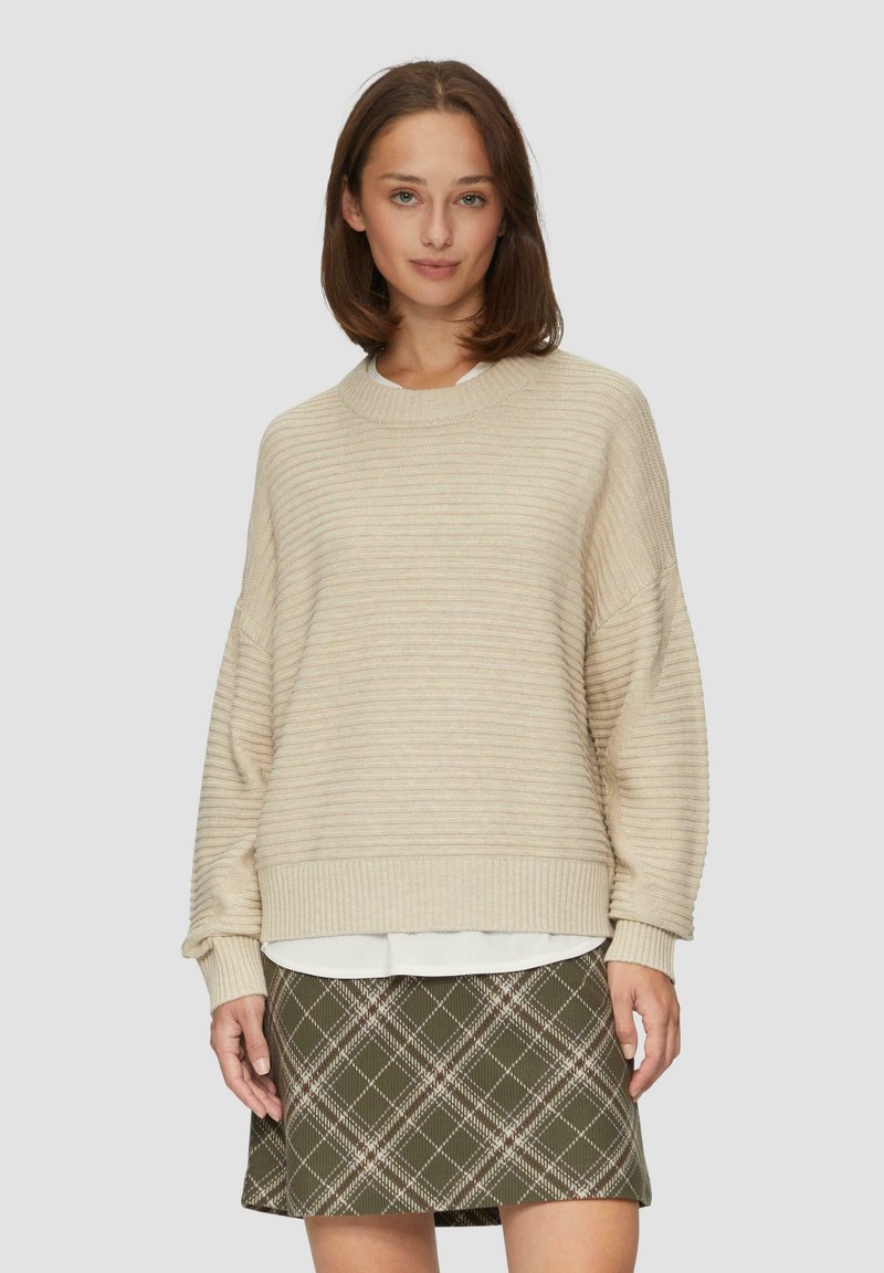 Beige gerippter Pullover mit rundem Halsausschnitt und überschnittenen Schultern, über einem weißen Hemd getragen, kombiniert mit einem grün karierten Rock.
