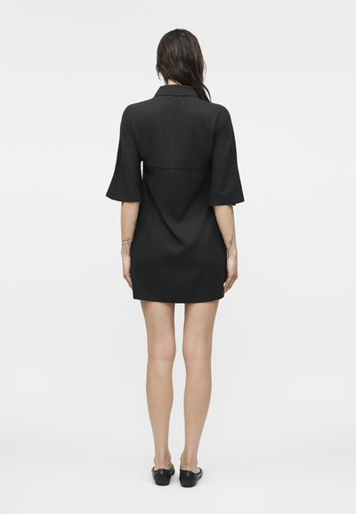 Γυναίκα που στέκεται με την πλάτη γυρισμένη, φοράει μαύρο φόρεμα με κοντά μανίκια και μαύρα φλατ παπούτσια, με ορατά τατουάζ στα χέρια της.