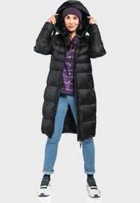 Langer schwarzer Daunenmantel mit Kapuze, mit einem gesteppten Design, kombiniert mit einem lila Shirt, blauen Jeans und sportlichen Turnschuhen.