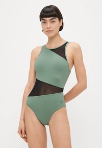 Ολόσωμο μαγιό σε ελιά πράσινο με μαύρες διαφανείς εισόδους. Διαθέτει ασύμμετρο σχέδιο, ψηλό λαιμό και σιλουέτα με υψηλή κοπή στα πόδια.