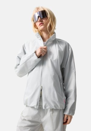 Jeune personne portant des lunettes de soleil réfléchissantes surdimensionnées et une veste grise claire à fermeture éclair avec un pantalon assorti, sur fond blanc.