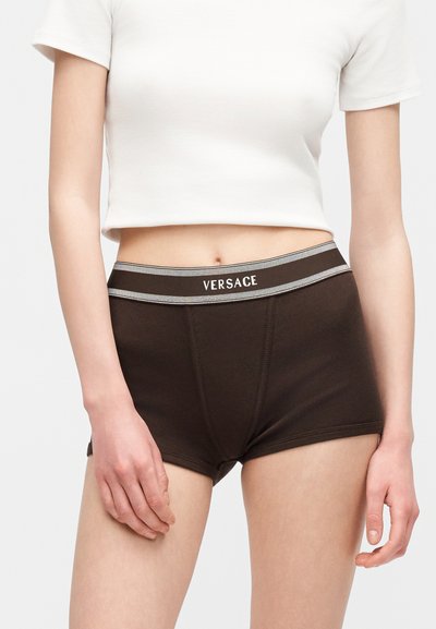 Femme portant un boxer brun foncé de la marque Versace et un top court blanc à manches courtes, debout devant un fond uni.