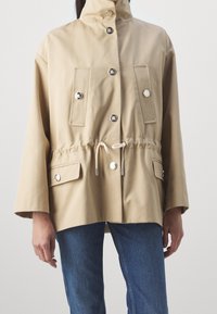 Chaqueta de algodón beige con cuello alto, botones de presión, cintura elástica y dos bolsillos frontales; cuenta con un cordón y mangas anchas.